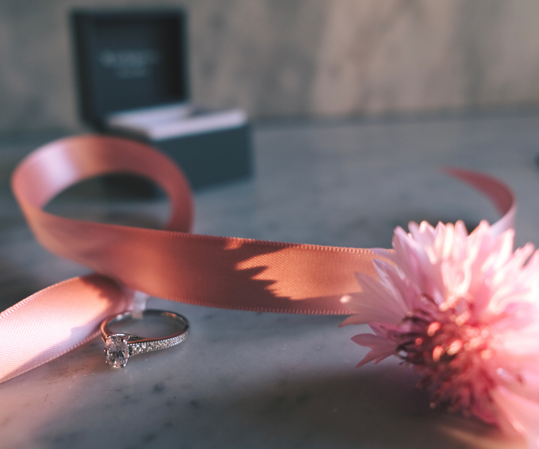Photograph of an engagement ring and an open ring box, with a pink ribbon and flower